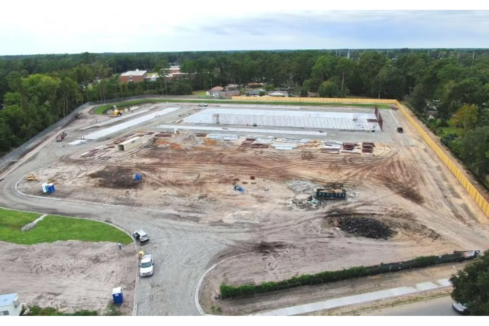 Aerial view of a large construction site with earthworks, concrete slabs, equipment, and surrounding trees under a cloudy sky