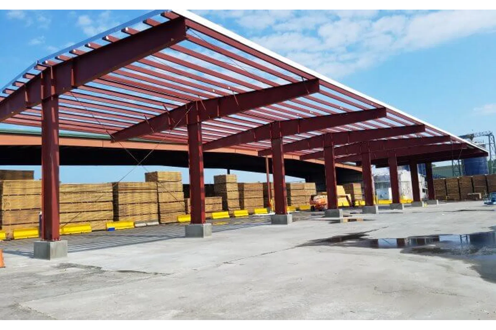 Red steel frame structure under construction in an outdoor lumber yard with stacked wooden planks and clear sky.