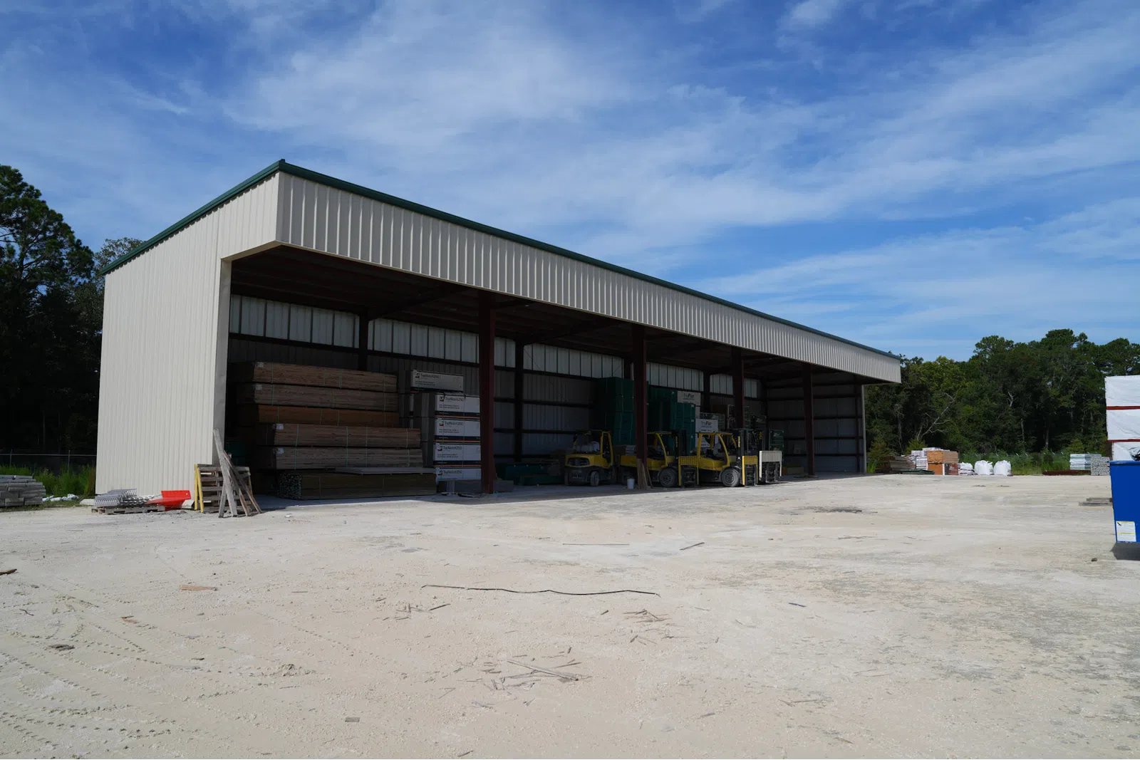 Large open metal storage shed housing construction materials and forklifts on a sunny day with clear blue sky.