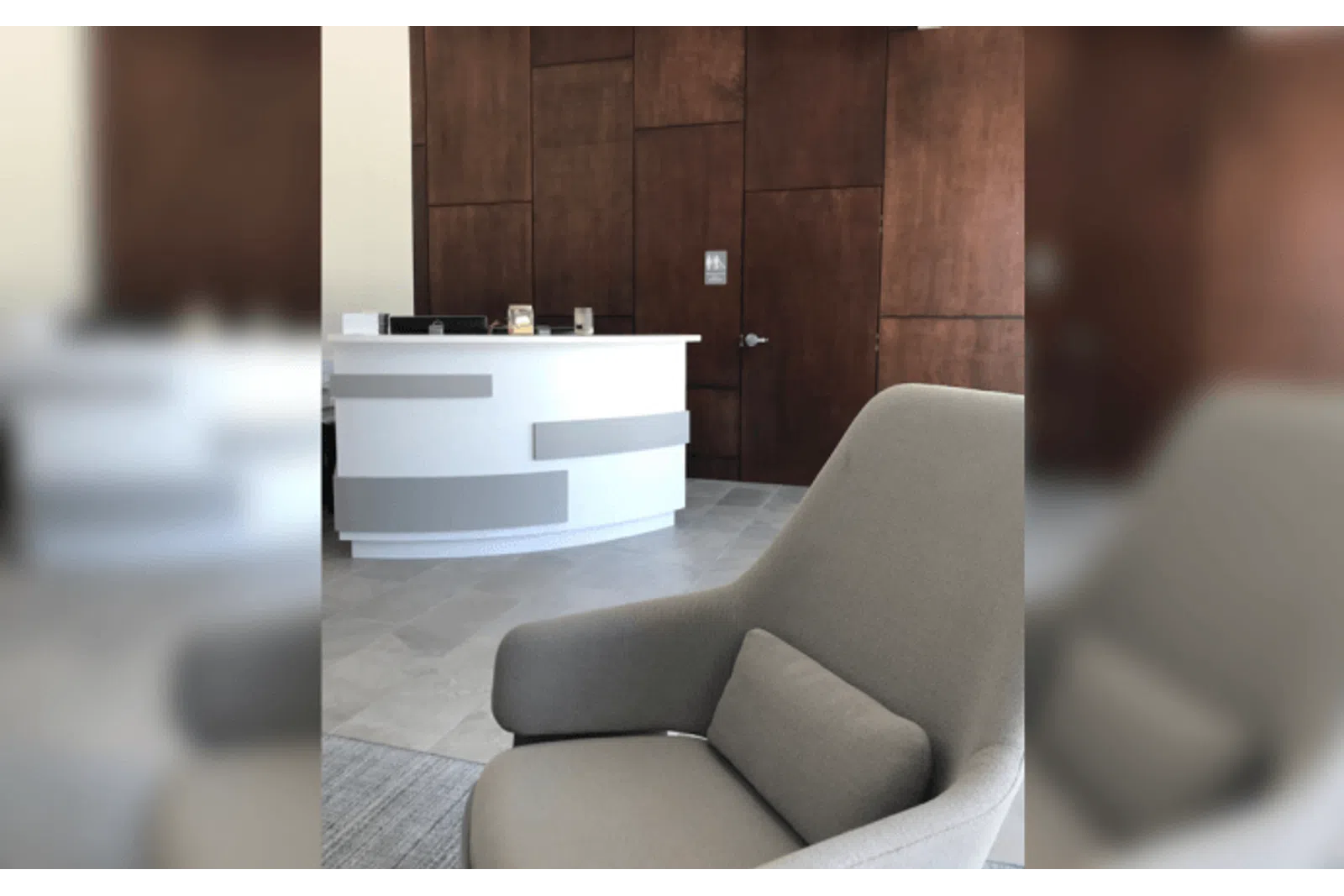 Modern reception area with a curved white desk, gray armchair, and wooden paneled wall with door.