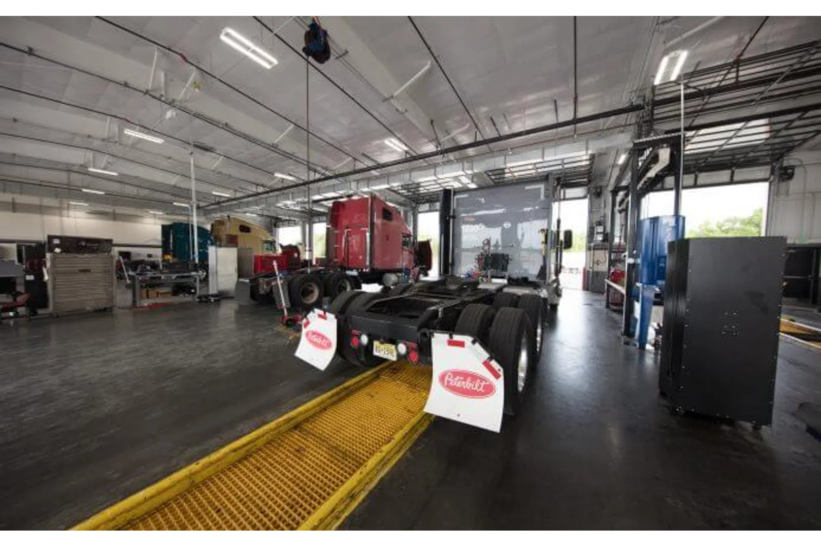 Inside a large truck repair shop with semi-trucks and maintenance equipment under bright lights.