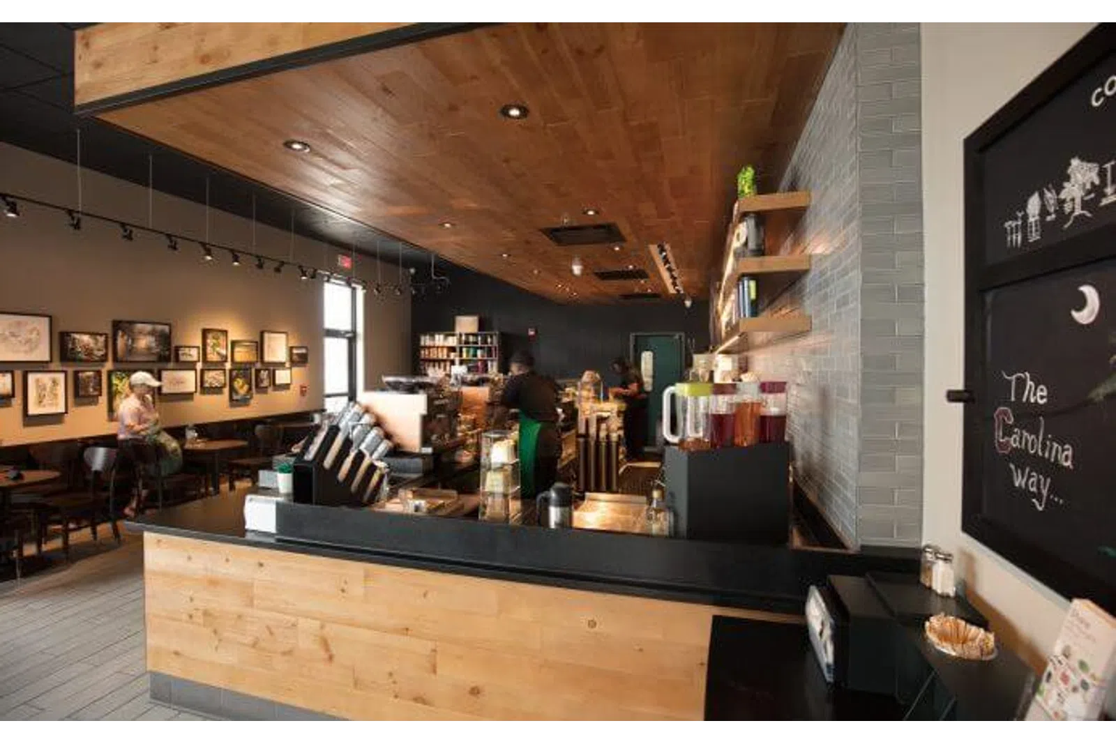 Cozy modern coffee shop interior with wooden accents, barista at counter, and customers seated by framed artwork.