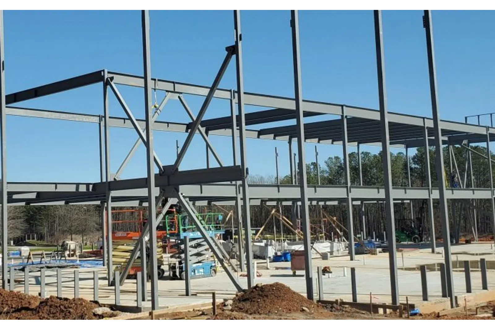 Steel framework of a building under construction on a clear sunny day with construction equipment and dirt piles.