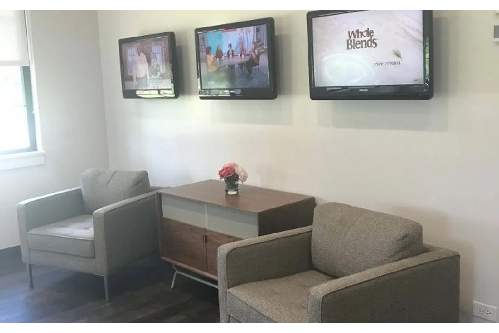 Modern waiting area with two gray armchairs, wooden cabinet, three wall-mounted TVs, and a small flower vase.