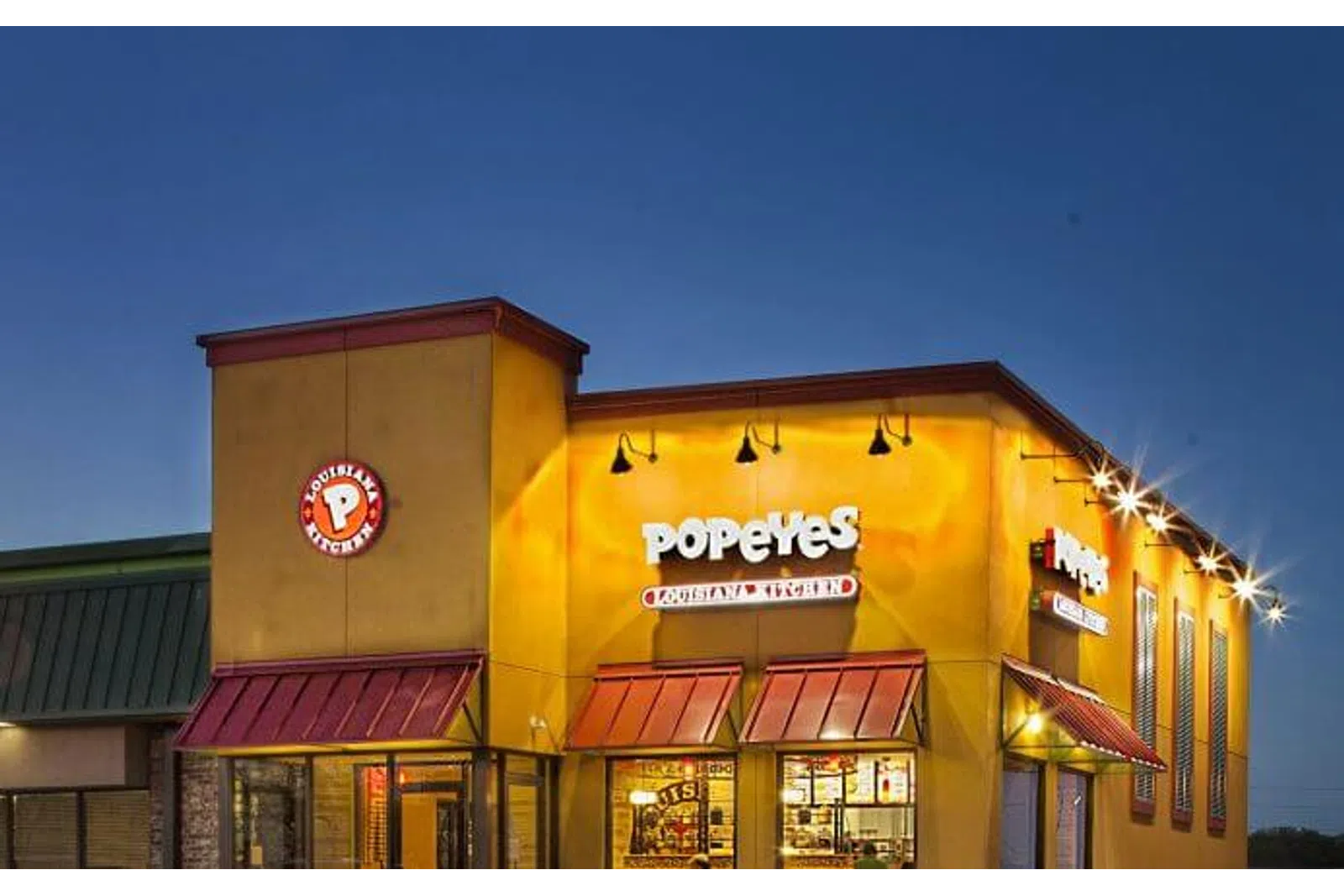 Popeyes Louisiana Kitchen restaurant building illuminated at dusk with logo and red awnings.