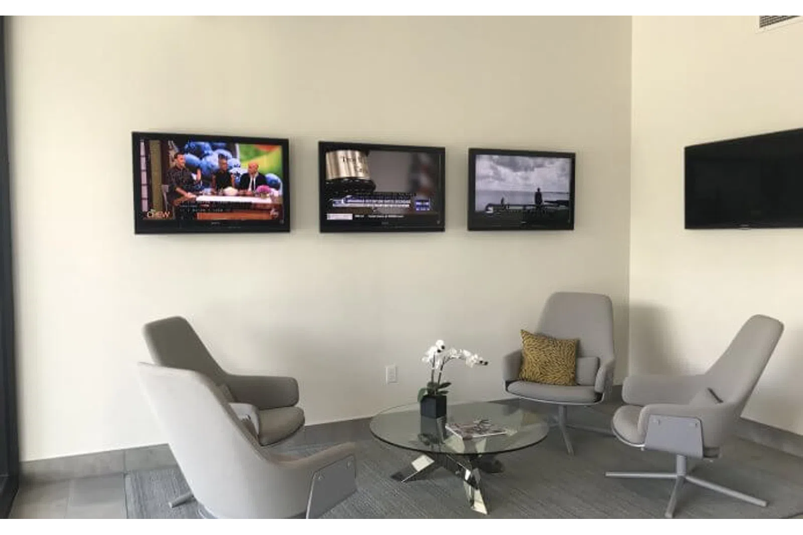 Modern waiting area with four gray chairs around a glass coffee table and three wall-mounted TVs.