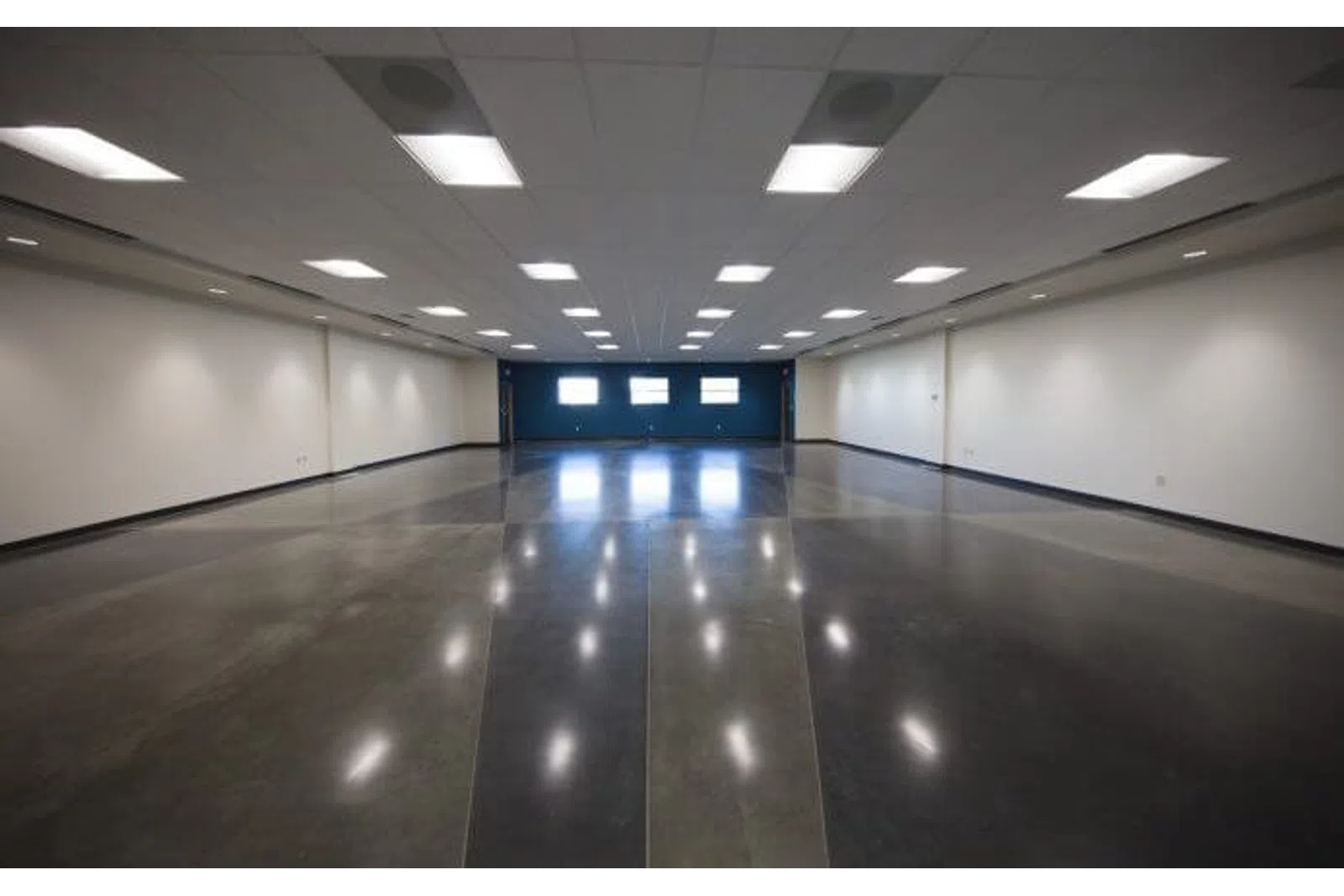 Spacious empty room with polished concrete floor, white walls, ceiling lights, and three windows at the far end.