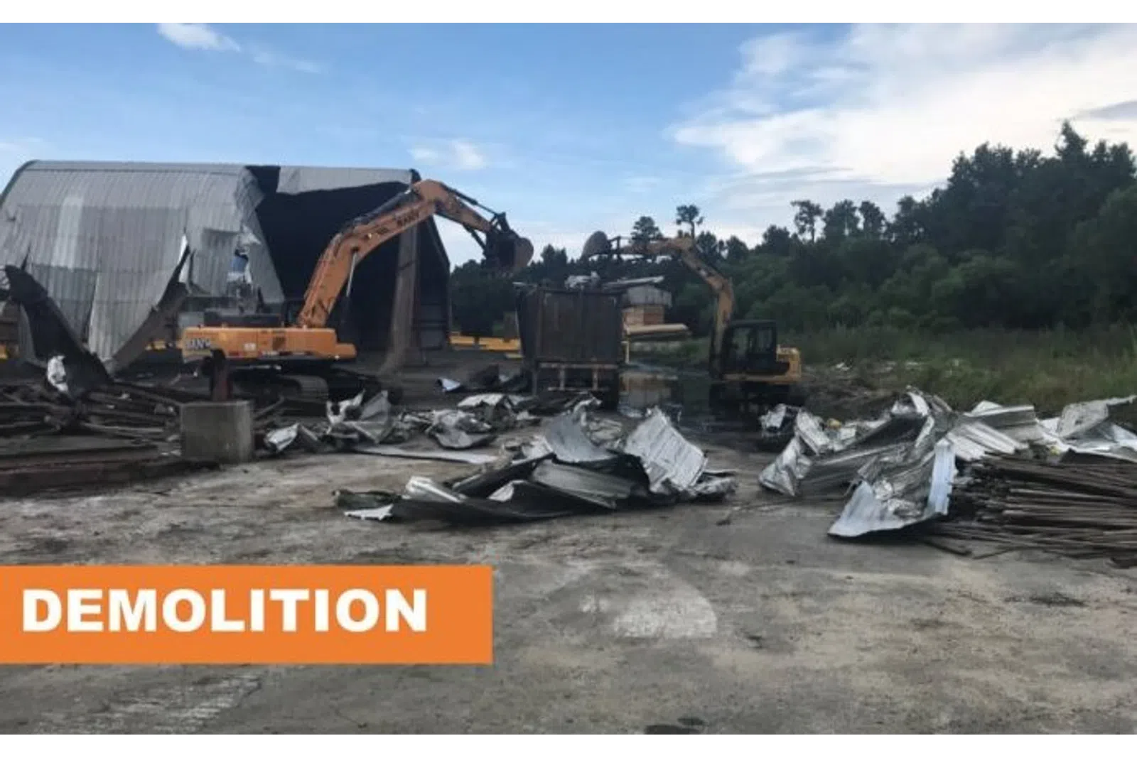 Two excavators demolishing a large metal building with debris scattered around on a construction site.