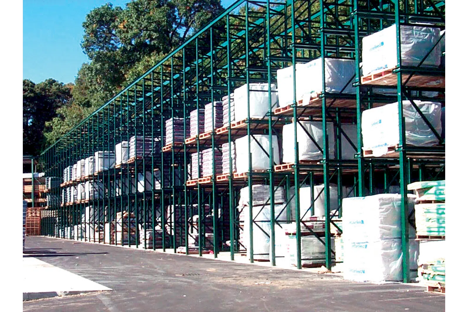Outdoor warehouse racks filled with stacked pallets wrapped in protective covers under clear sky.