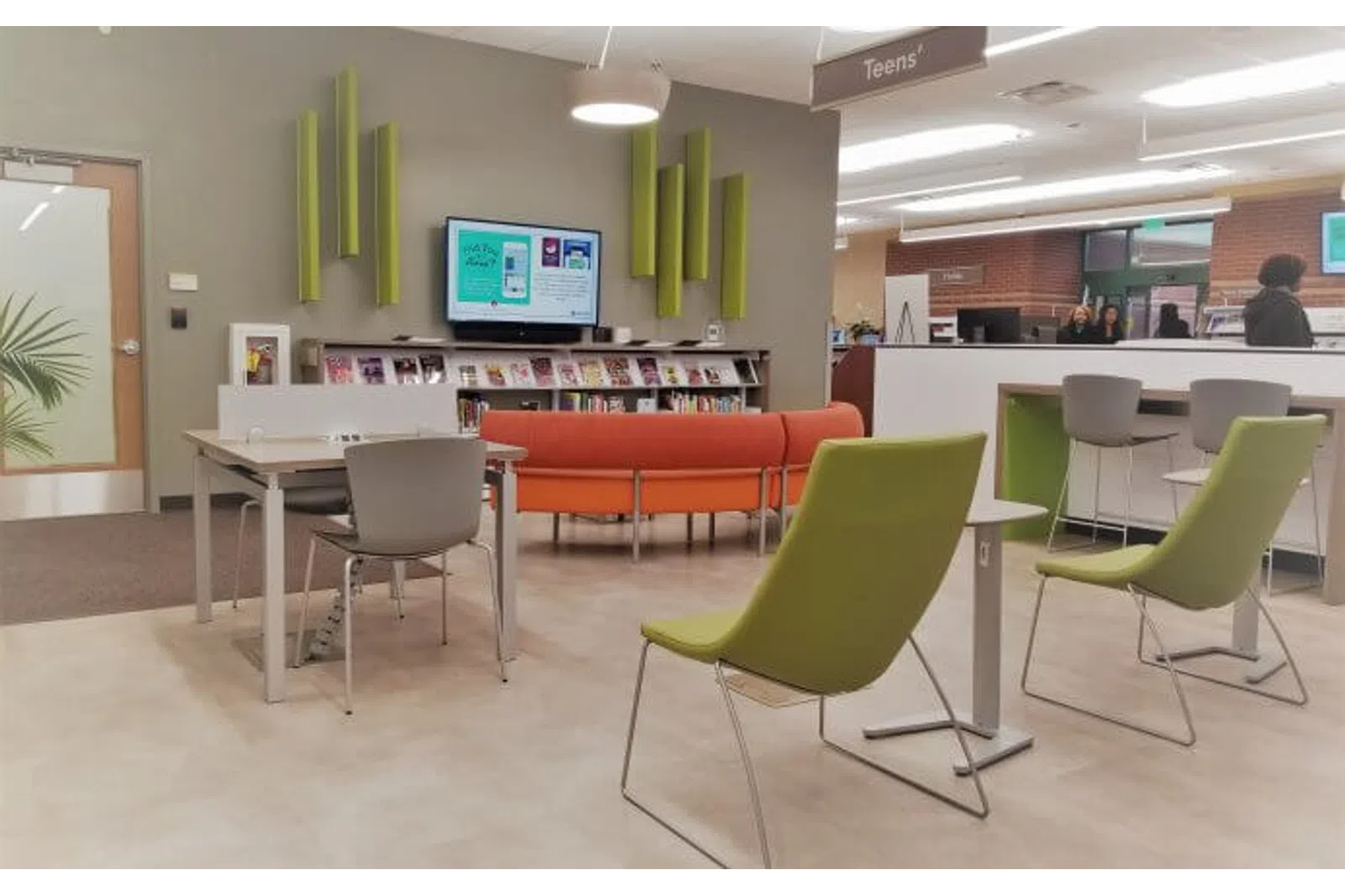 Modern library teen area with green chairs, orange couch, tables, bookshelves, and digital information screen.