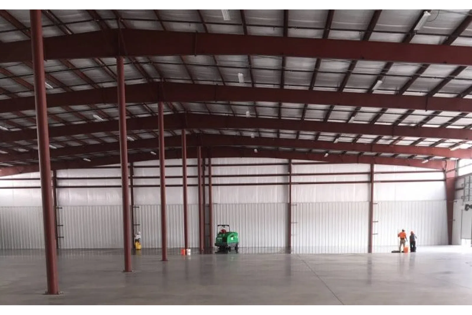 Large empty warehouse interior with steel beams, concrete floor, and two workers operating cleaning equipment.