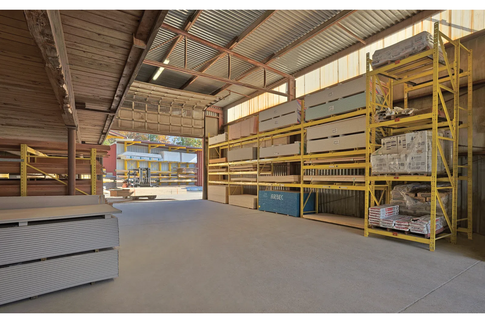 Warehouse interior with construction materials stored on yellow metal shelves and pallets on concrete floor.