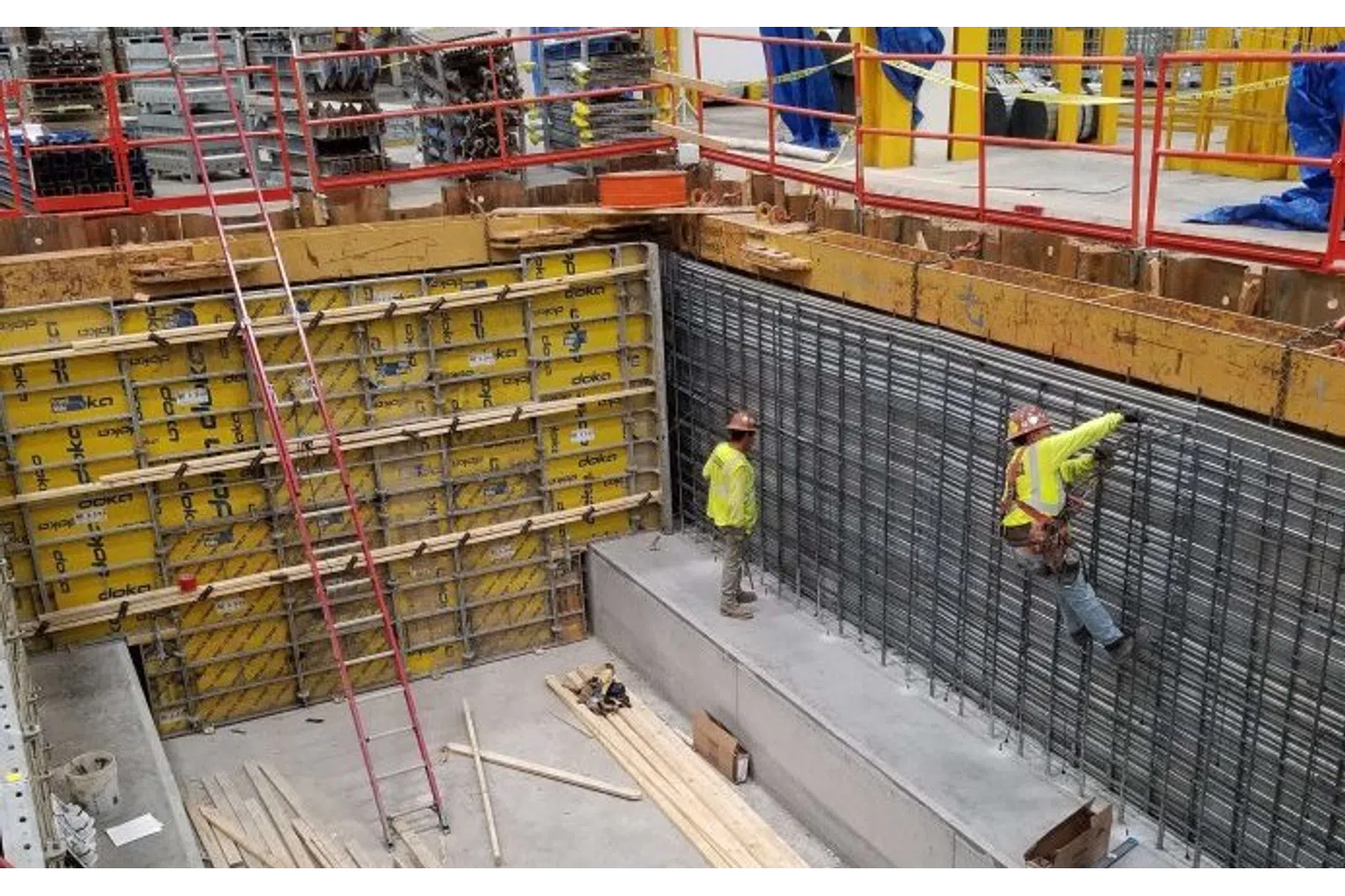 Construction workers in safety gear installing steel reinforcement inside a concrete foundation with yellow formwork panels.