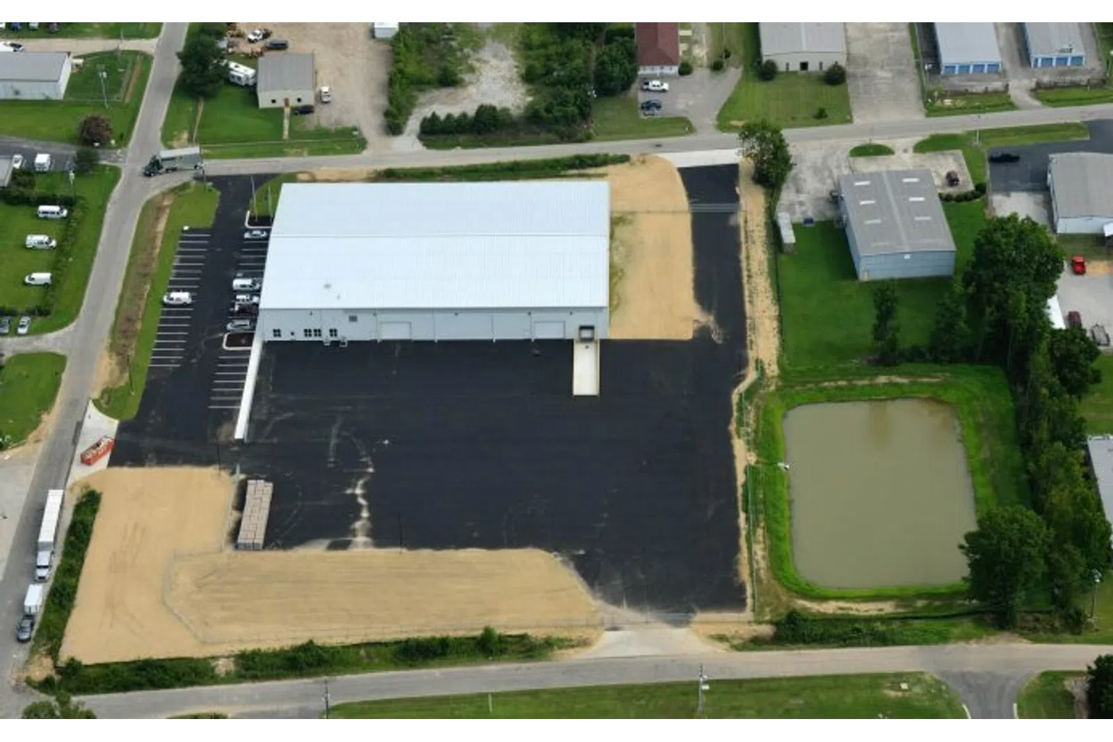 Aerial view of industrial warehouse with large parking lot and adjacent pond surrounded by greenery and roads.