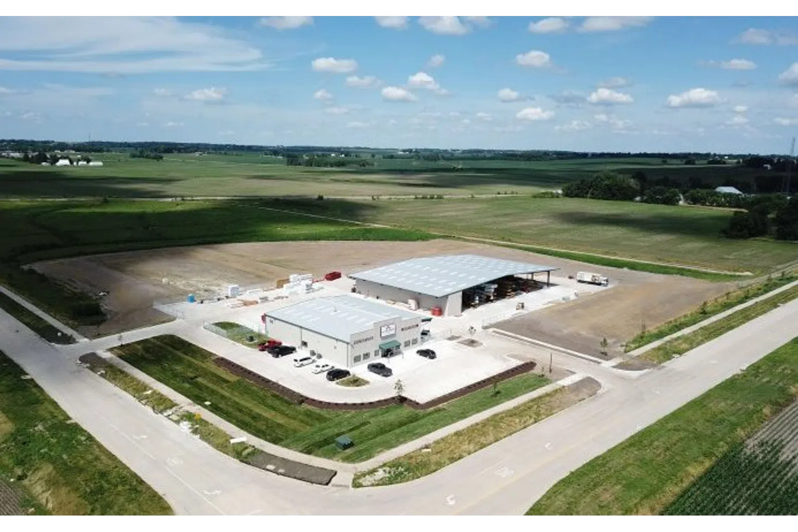 Aerial view of industrial warehouse buildings surrounded by farmland and roads under a blue sky with scattered clouds