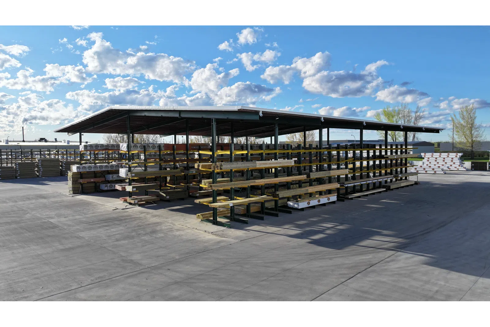 Outdoor lumber storage with organized metal racks under a metal roof on a sunny day with scattered clouds.