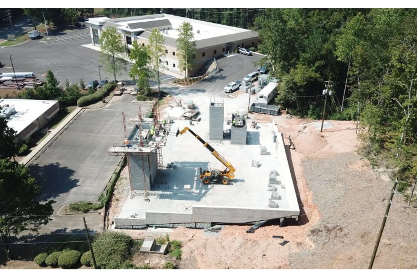 Aerial view of a construction site with concrete foundation, crane, and workers amidst surrounding trees and buildings.