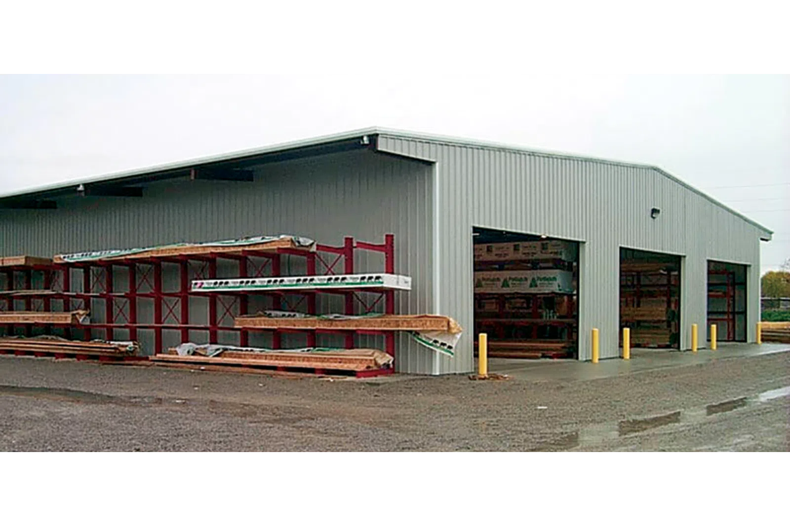 Exterior view of a metal storage building with open bays and lumber racks on a gravel lot