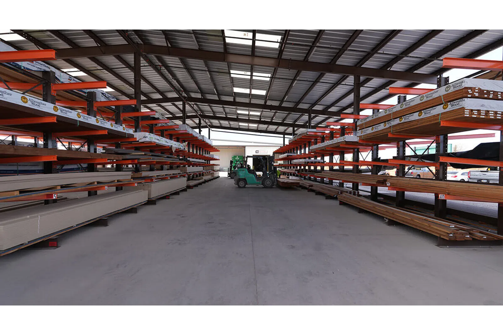 Warehouse interior with metal and wood materials stored on orange racks and forklifts in the center aisle.
