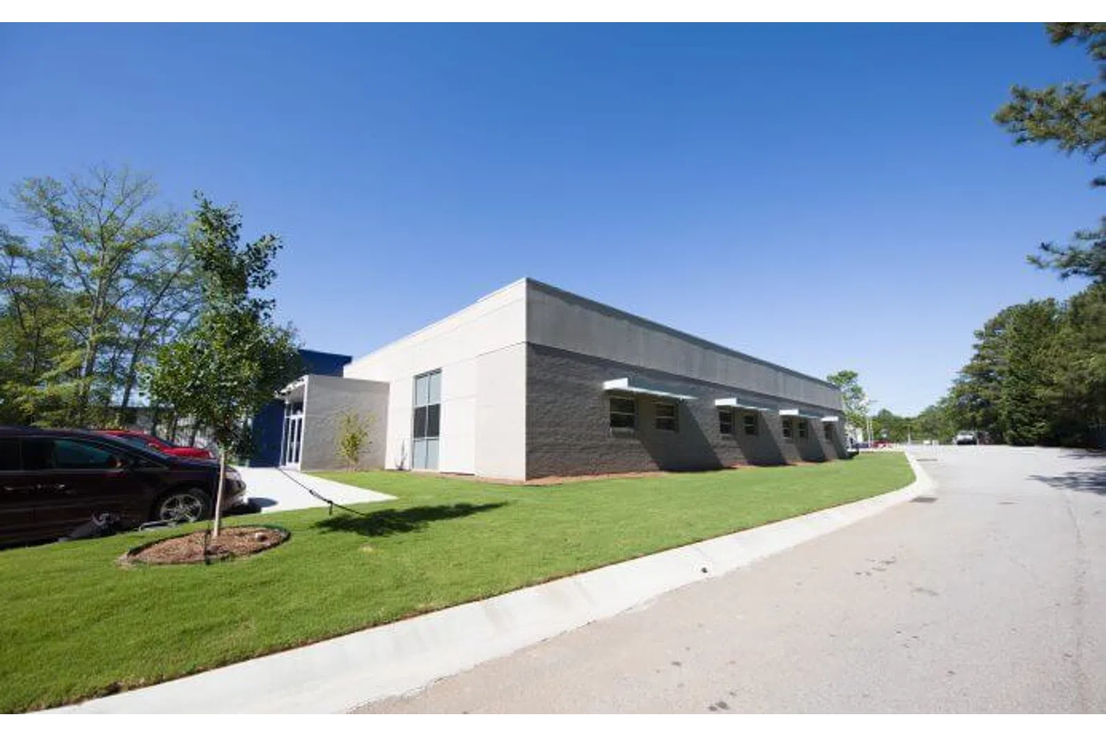 Modern single-story commercial building with gray walls, green lawn, parked cars, and clear blue sky.
