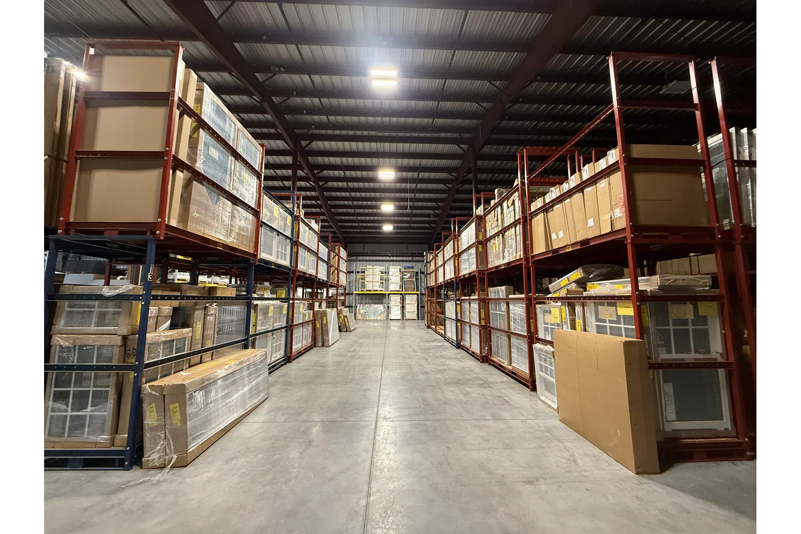 Spacious warehouse aisle with tall red metal racks filled with cardboard boxes and packaged goods under bright ceiling lights