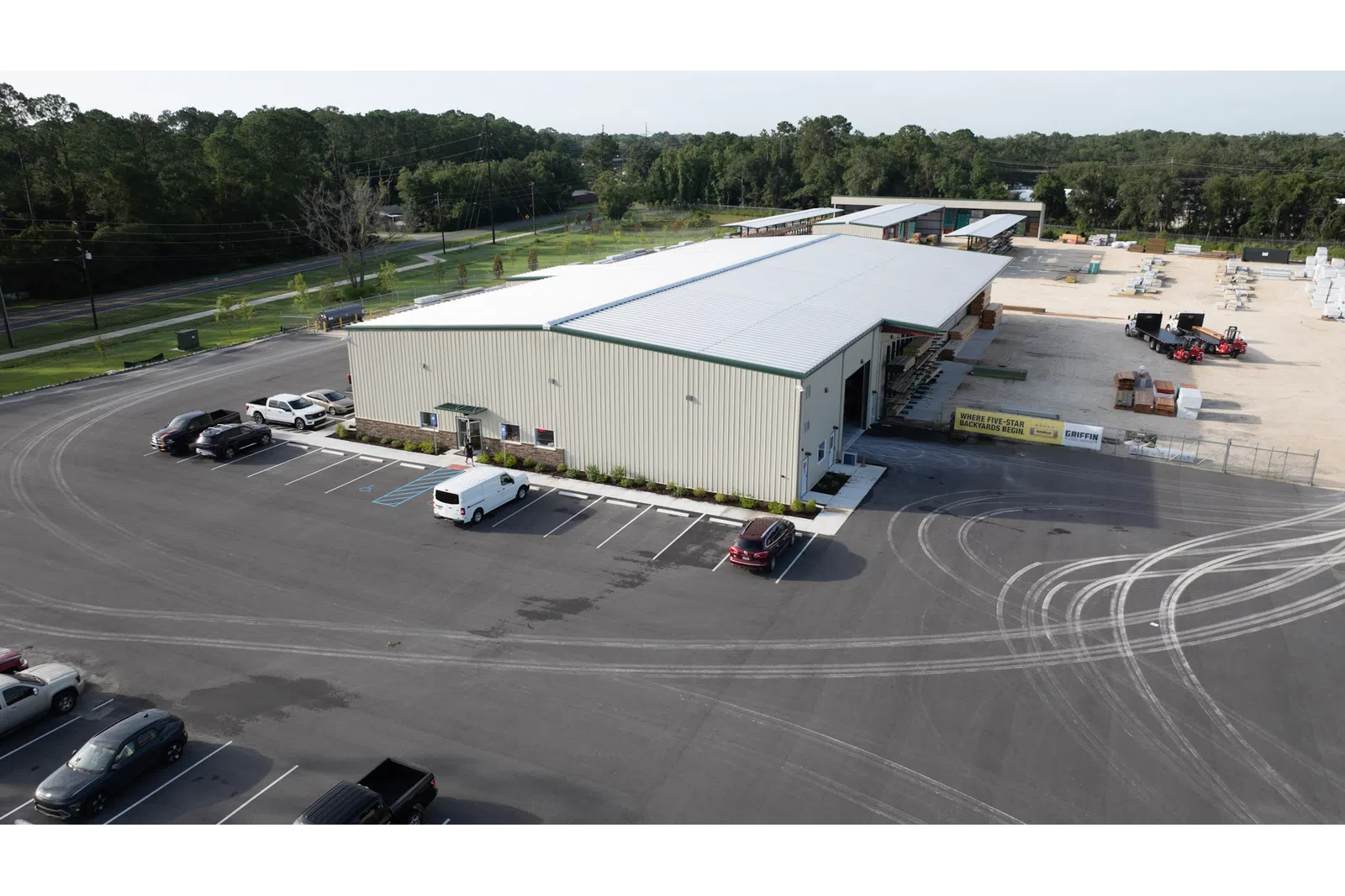 A large industrial warehouse with several parked vehicles and tire marks in a spacious asphalt lot surrounded by trees.