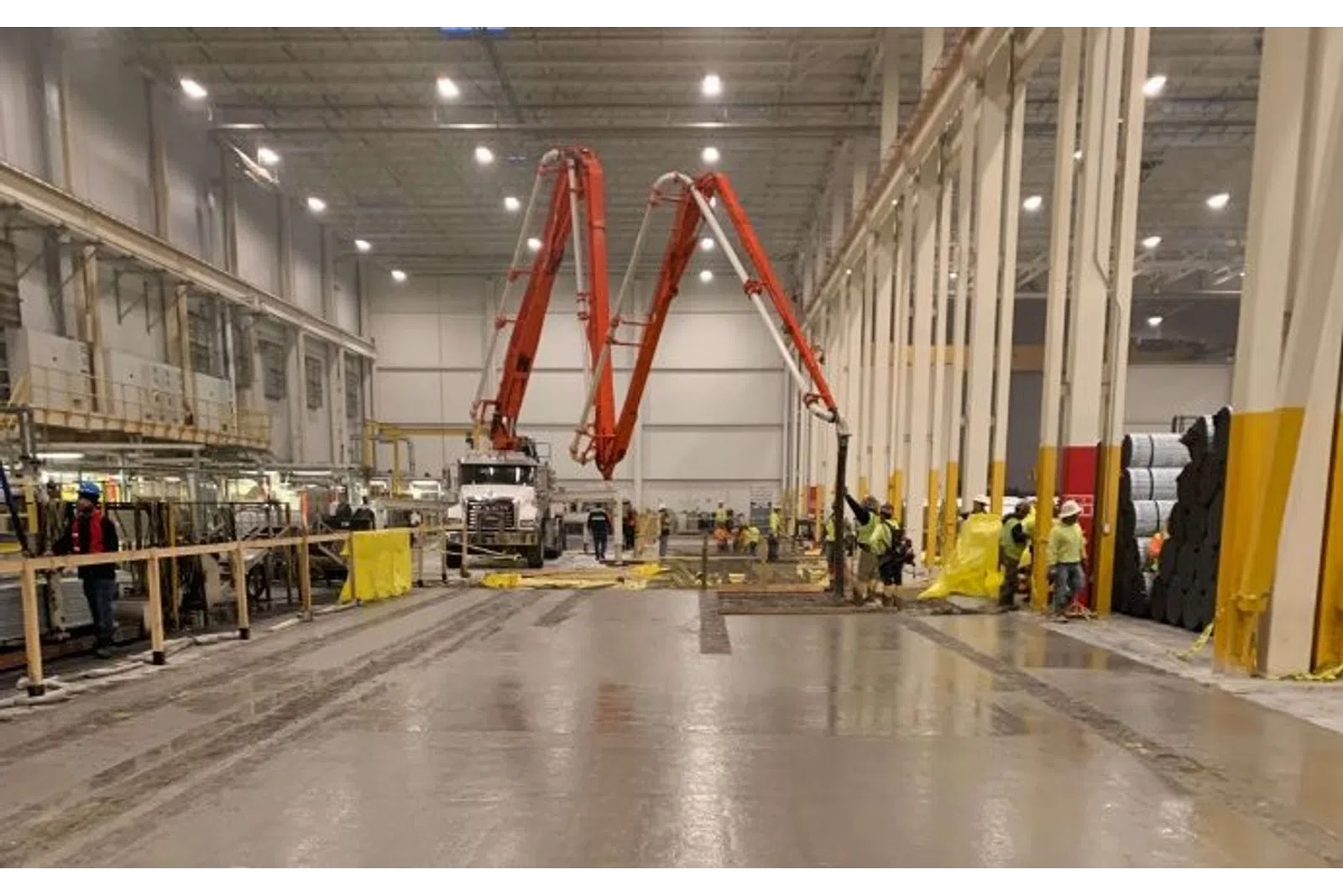 Workers using concrete pump trucks inside a large industrial warehouse with high ceilings and pillars.