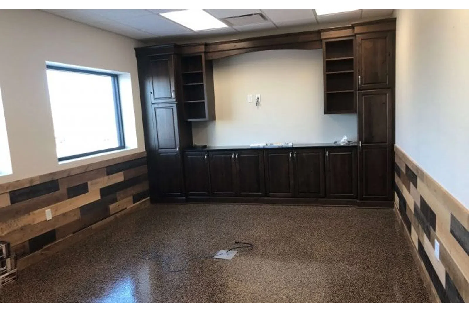 Empty room with polished concrete floor, wood panel walls, large window, and dark wooden cabinets against the wall.