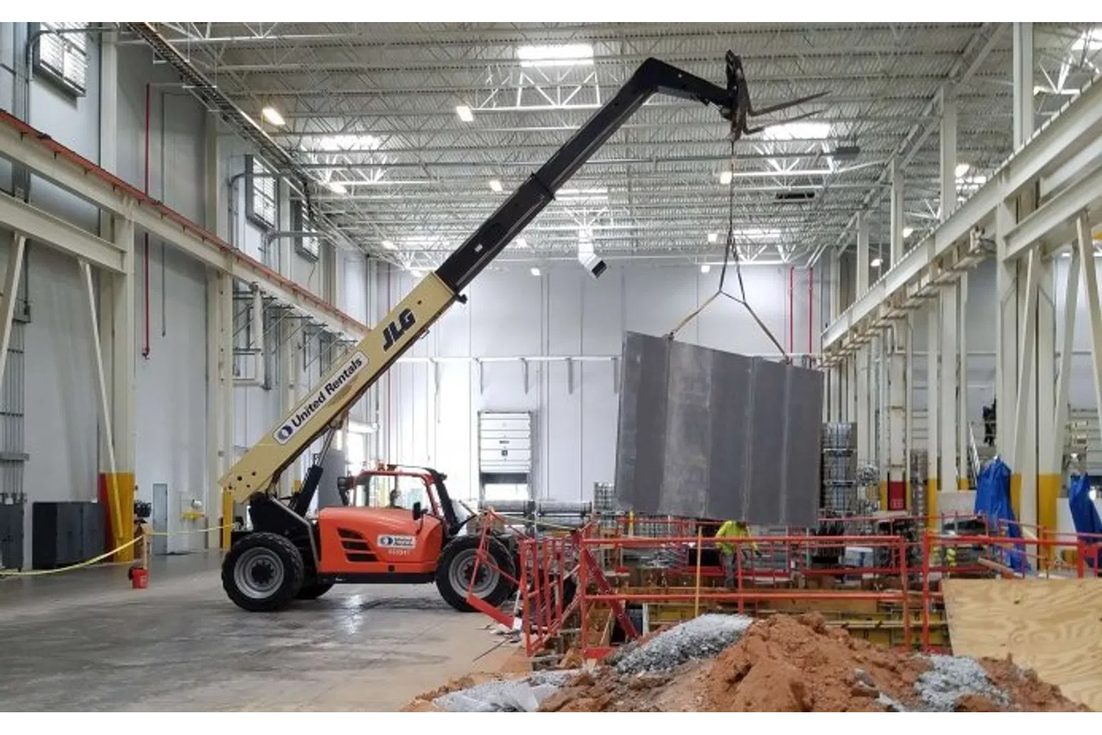 Orange JLG telehandler lifting a large metal panel inside a spacious industrial warehouse under construction.