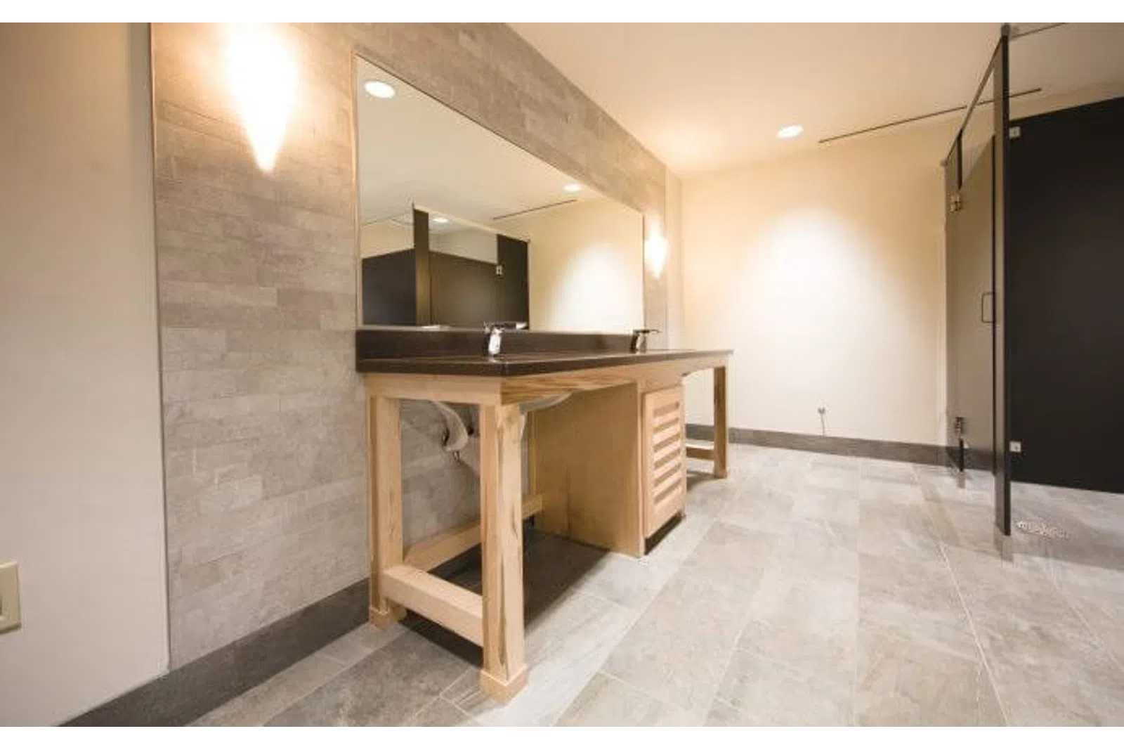 Modern public restroom with wooden vanity, large mirror, tiled floor, and black partition stalls under bright lighting