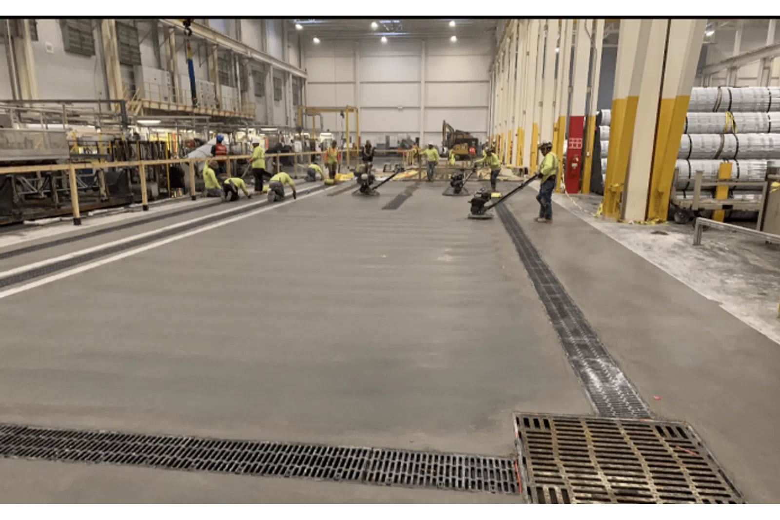 Workers smoothing and finishing a large concrete floor inside an industrial warehouse with heavy machinery.
