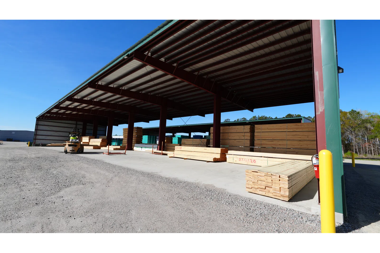 Outdoor lumber storage area with stacks of wooden boards under large metal shelter on sunny day.