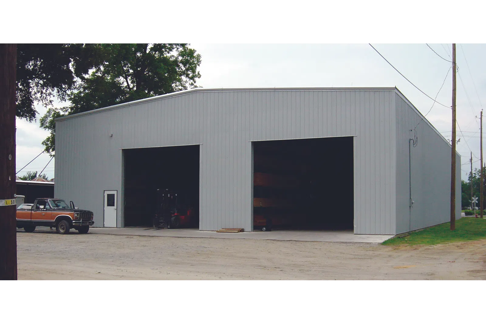 Large gray metal warehouse building with two open garage doors and a parked vintage truck outside under trees.