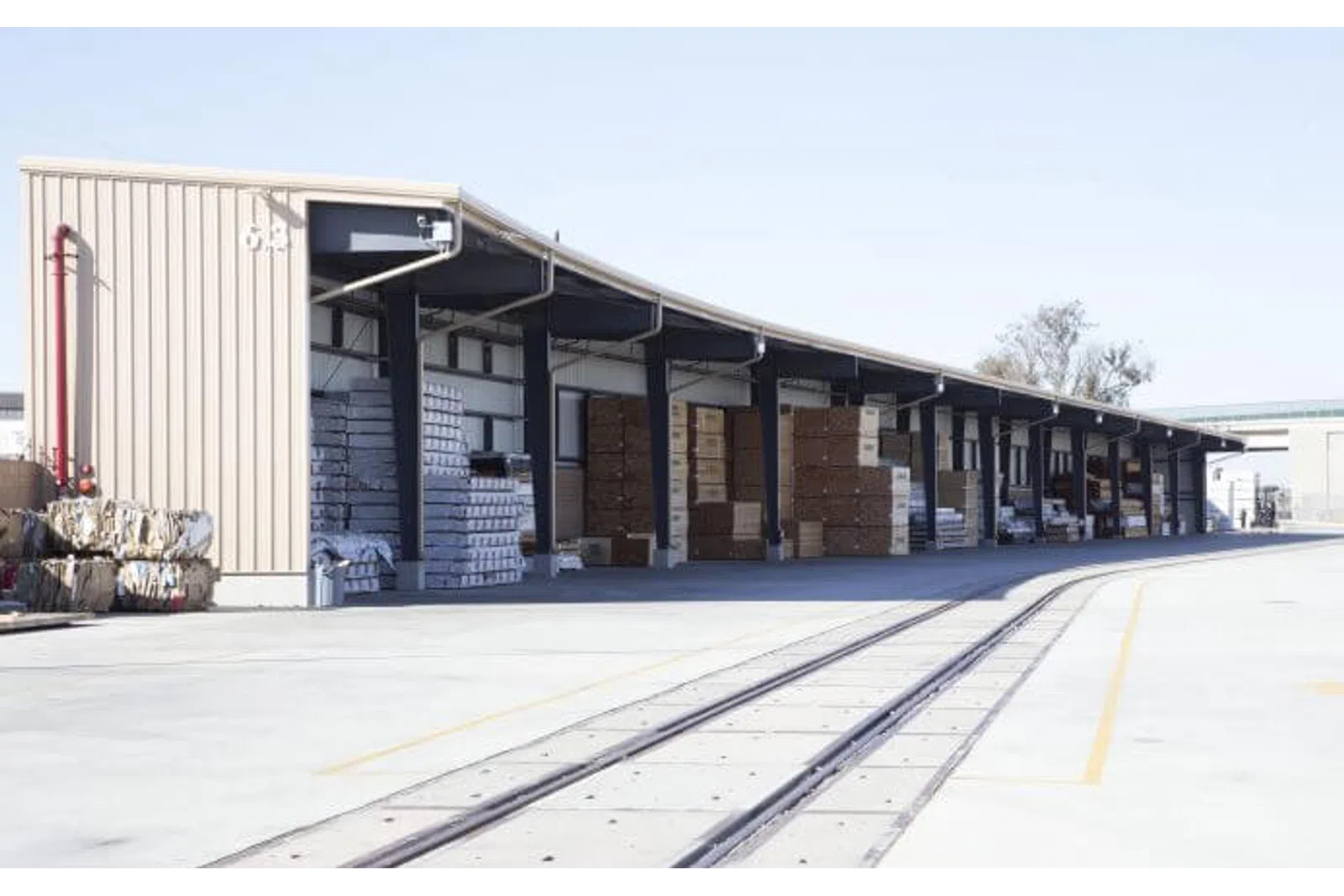 Outdoor warehouse with stacked boxes and pallets under a long covered loading dock near railway tracks.
