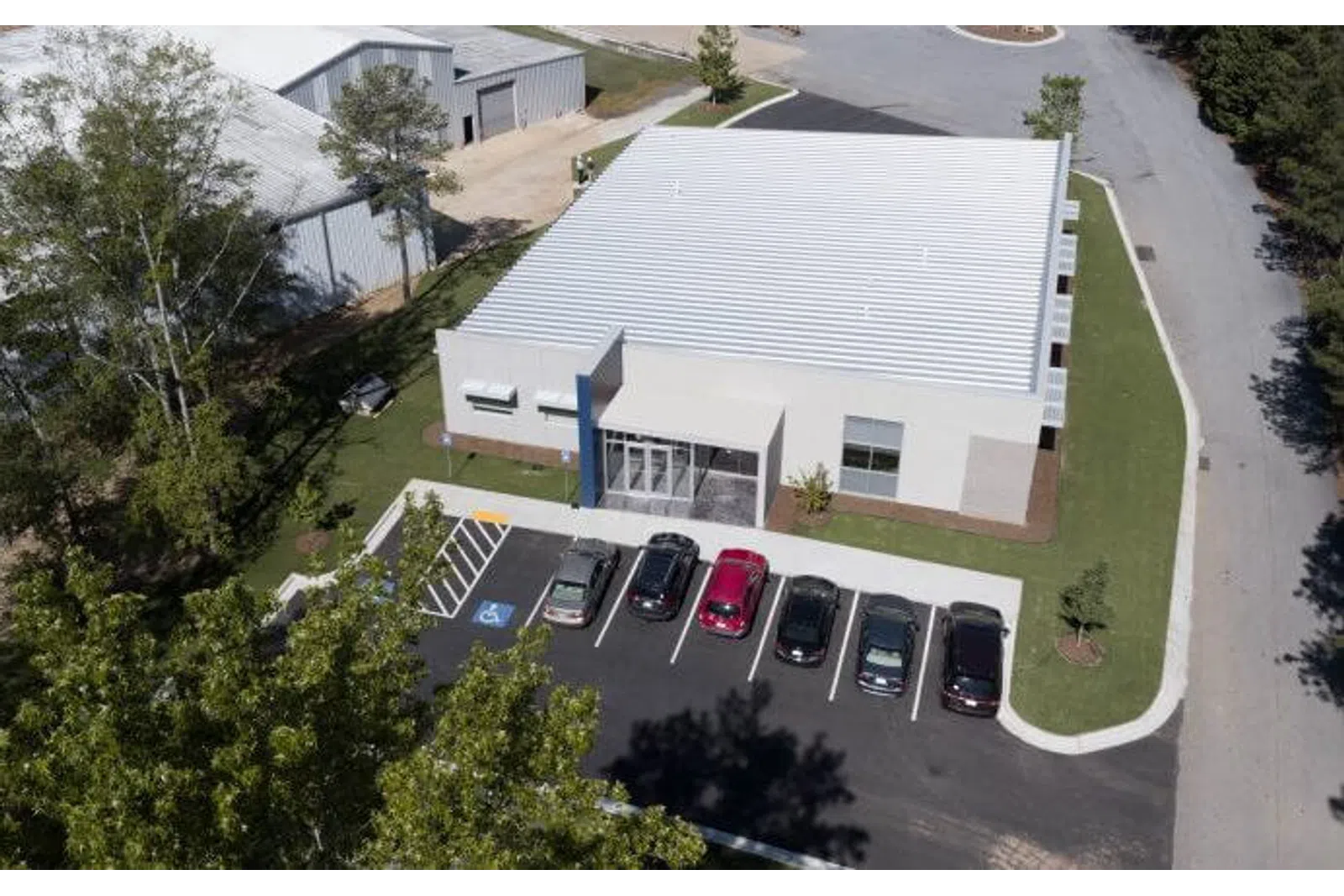Aerial view of a modern commercial building with a parking lot and six parked cars surrounded by trees and a road.