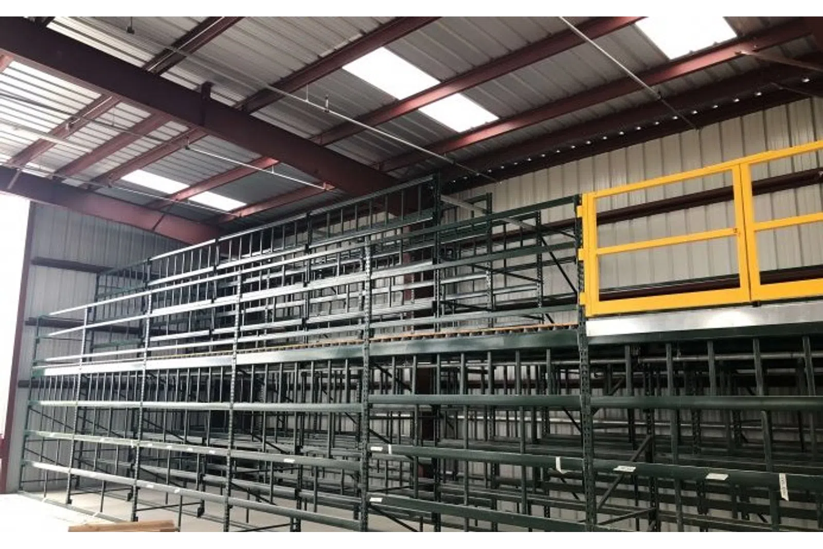 Empty metal storage racks inside a warehouse with a yellow safety gate on the upper platform under a skylit roof.