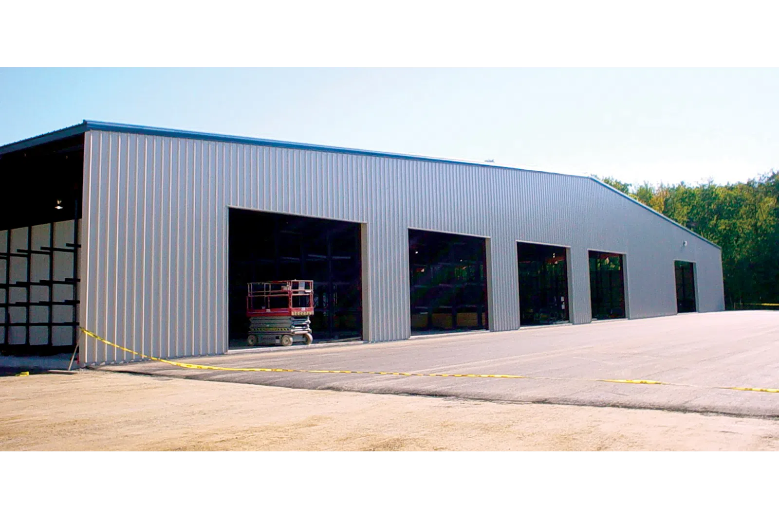 Large metal warehouse with multiple open bays under clear sky and surrounding green trees.