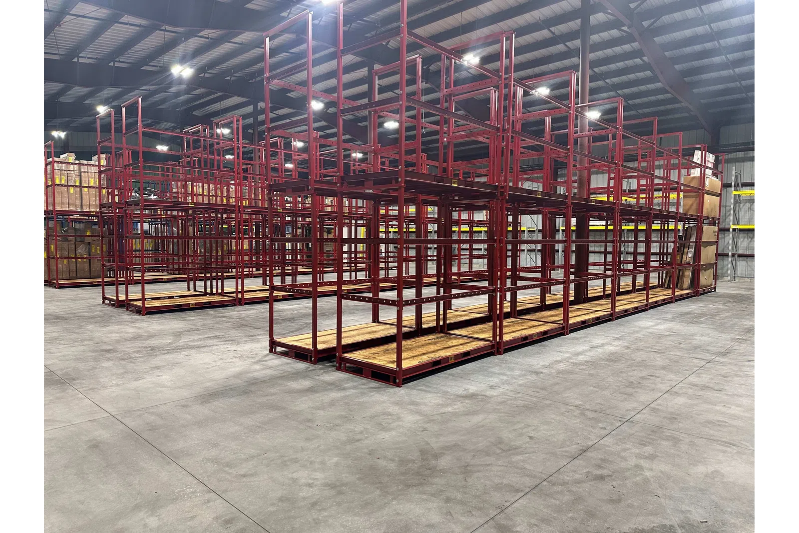 Empty red metal storage racks with wooden shelves inside a large warehouse with concrete floors and steel ceiling.