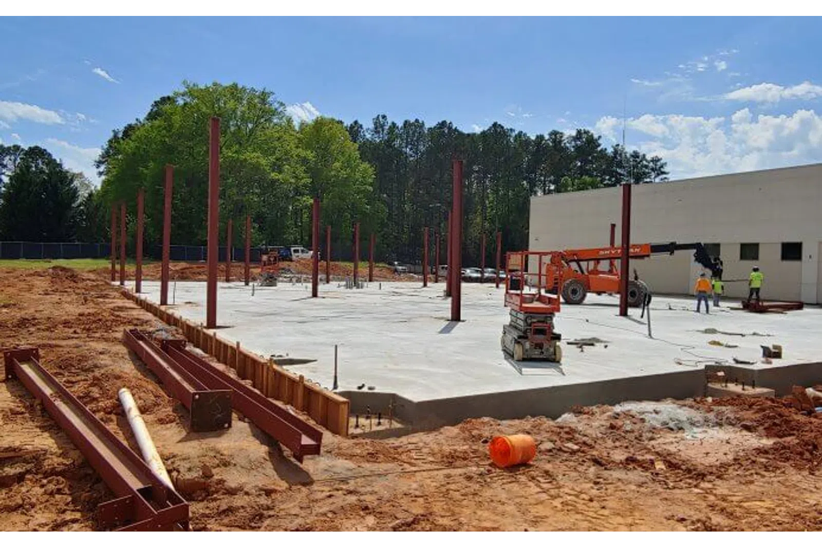 Construction site with steel beams and concrete foundation amid trees and workers under blue sky.
