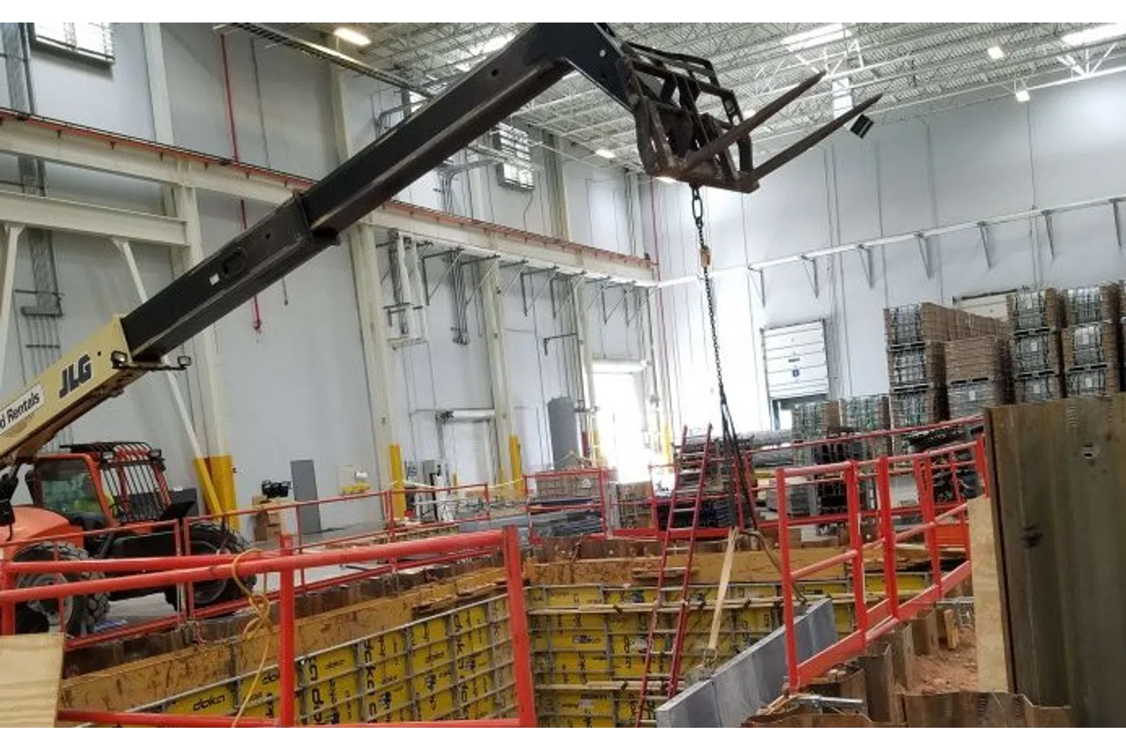 Indoor construction site with a crane lifting materials over a foundation pit surrounded by safety railings.