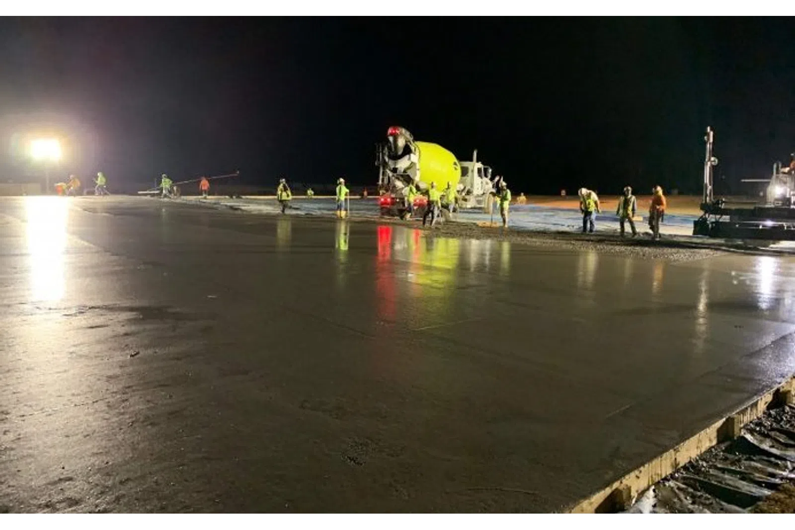 Nighttime construction crew pours and smooths concrete slab under bright lights with cement mixer truck present.