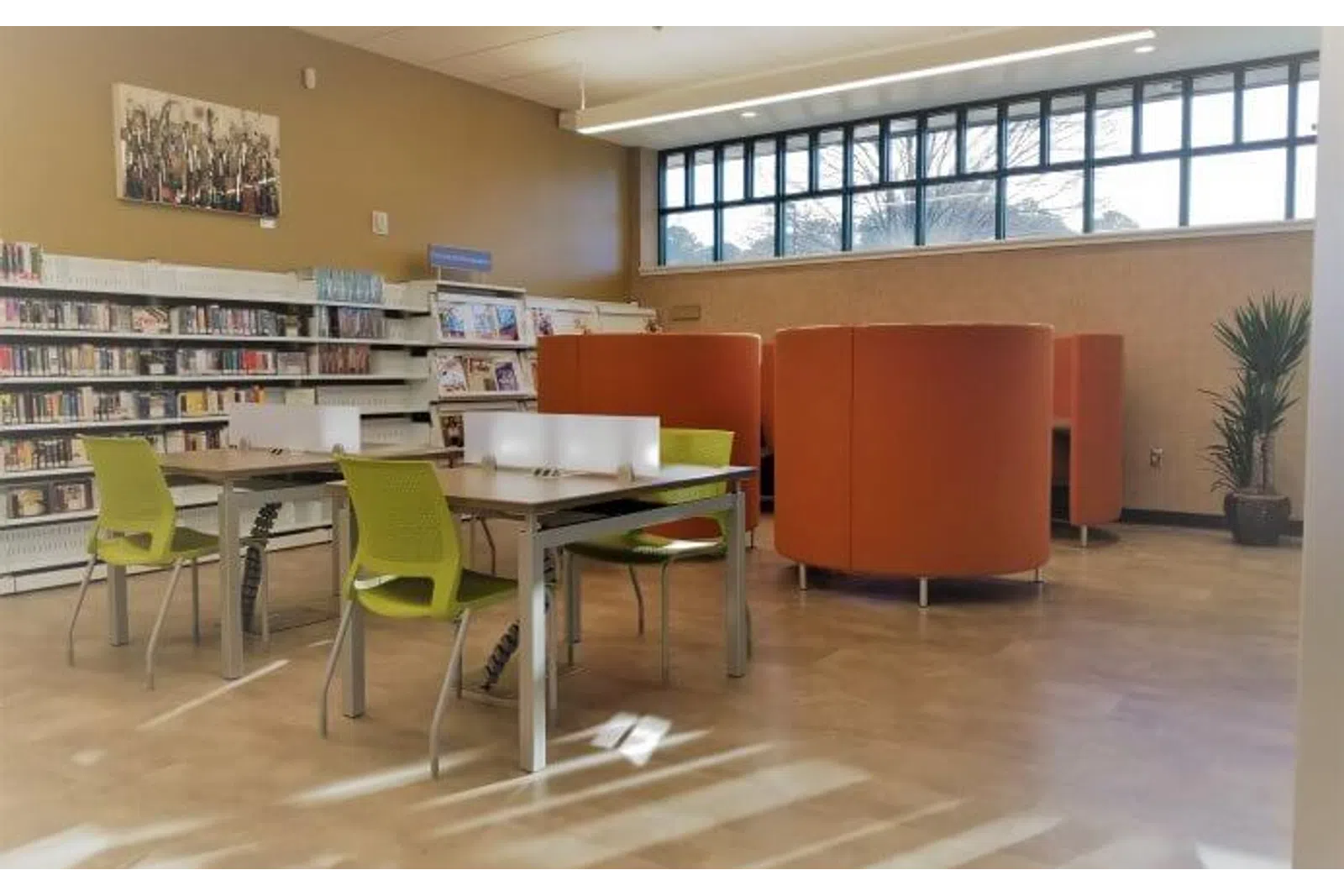 Modern library interior with green chairs, tables, orange curved booths, bookshelves, and large windows letting in natural light