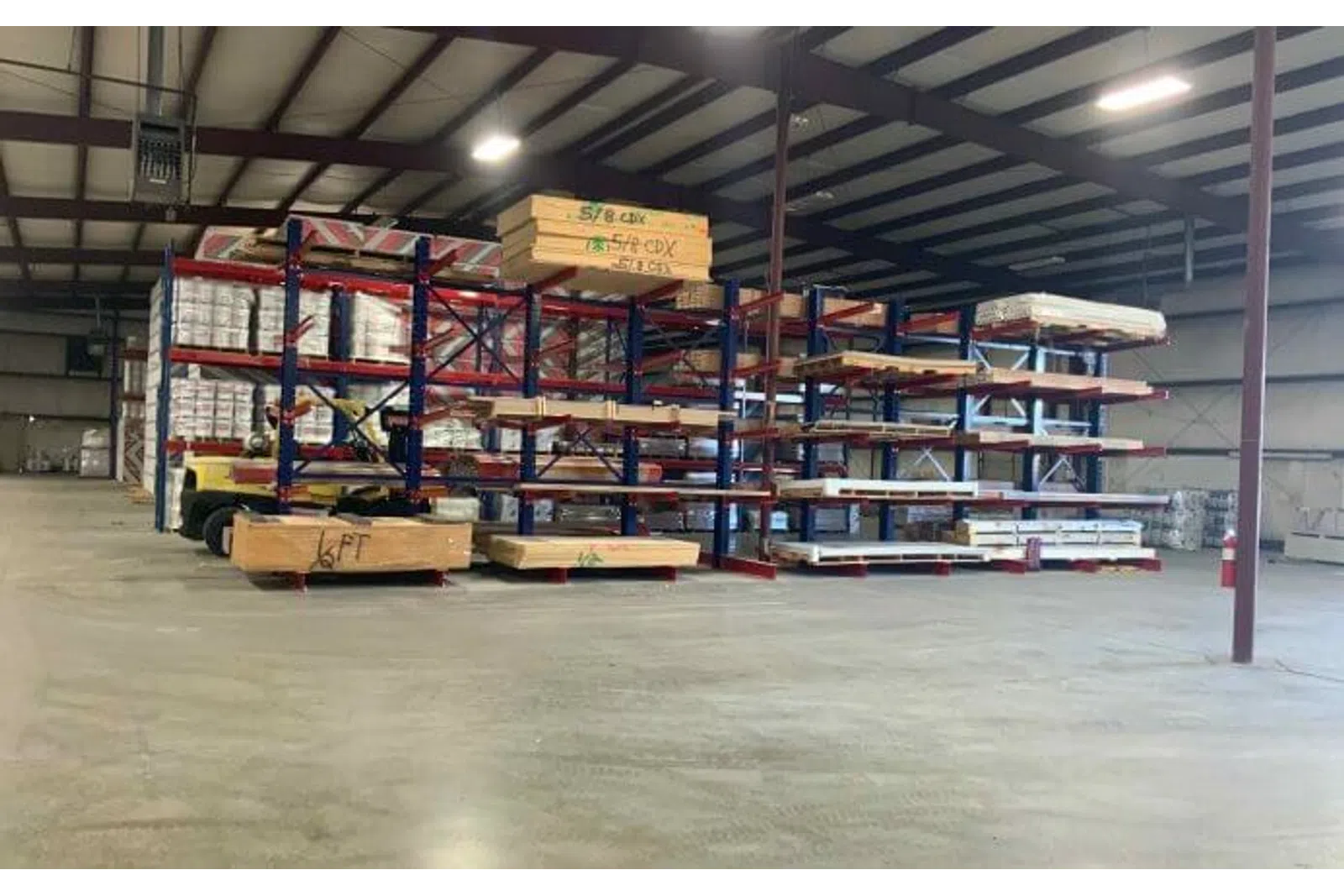 Warehouse interior with steel racks holding wooden planks and plywood sheets under fluorescent lights.