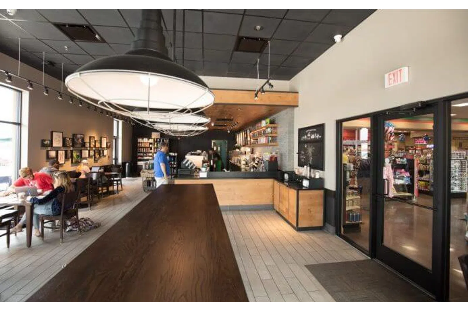Modern coffee shop interior with wooden tables, large pendant lights, and customers sitting near windows.