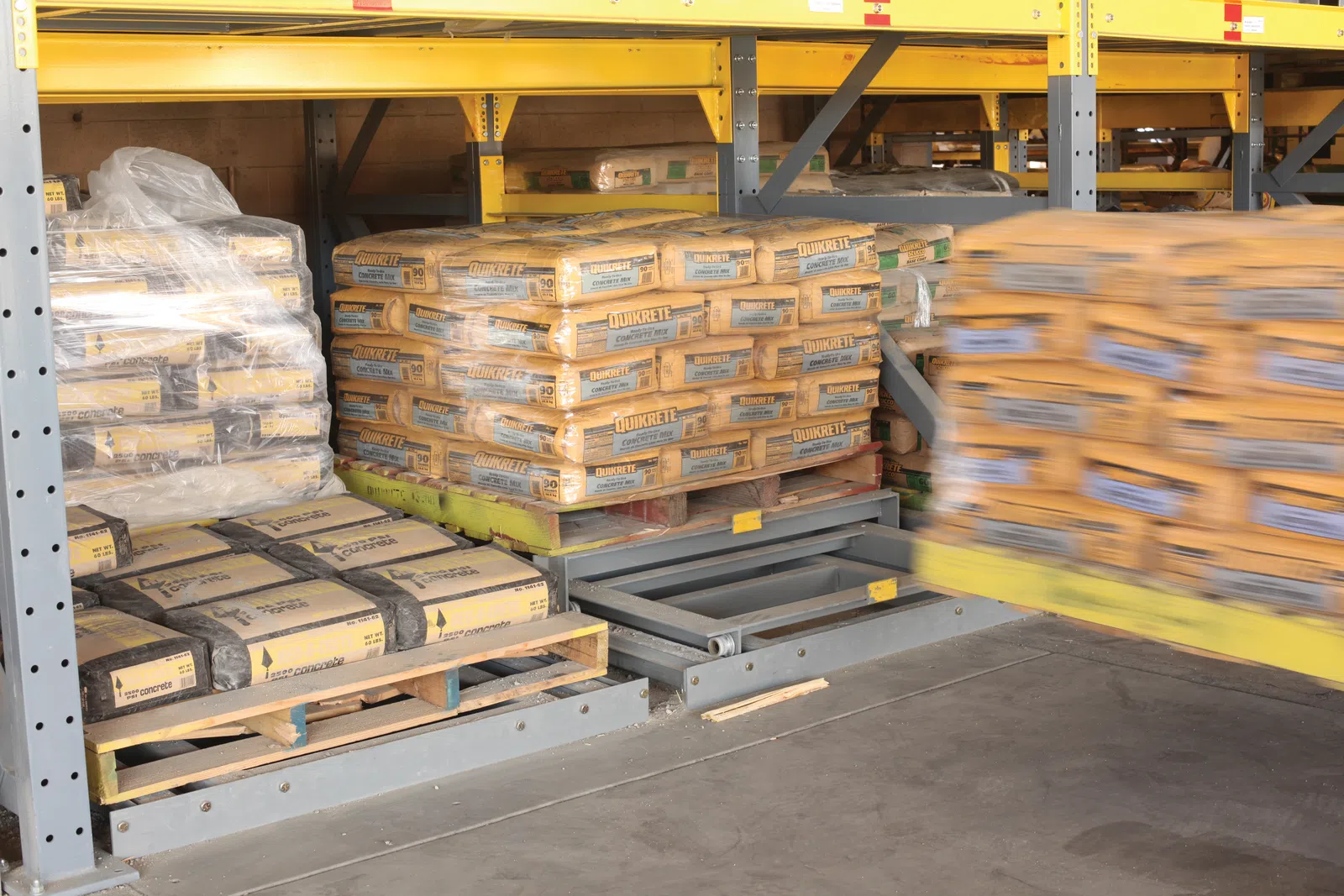 Pallets of Quikrete bags stacked on metal shelves in a warehouse with motion blur on one side.
