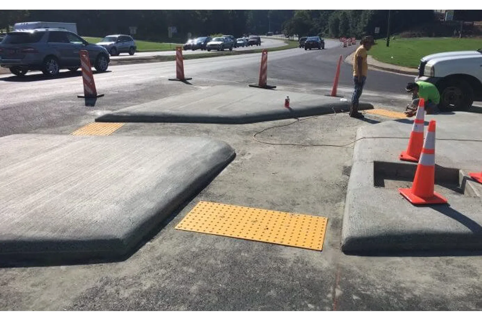 Newly constructed concrete curb ramps with tactile paving and traffic cones near a busy road under clear sky.