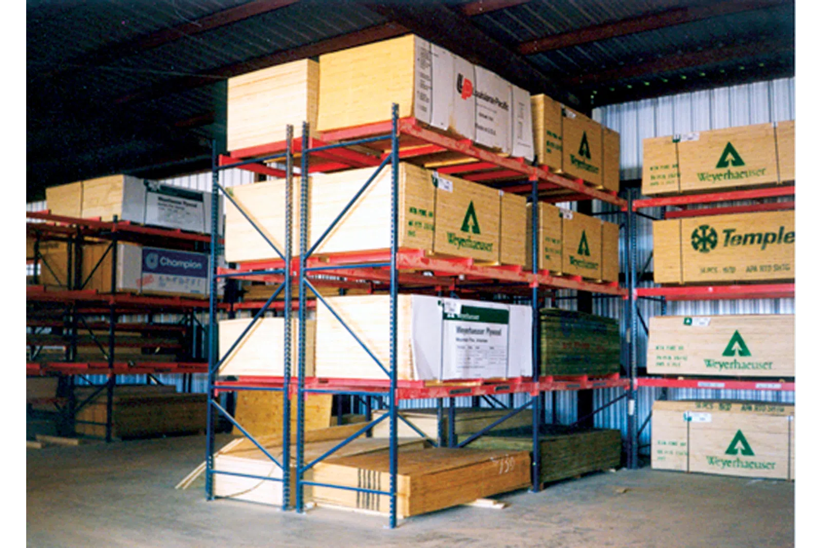 Warehouse interior showing stacks of lumber on metal racks under a corrugated roof with branded packaging