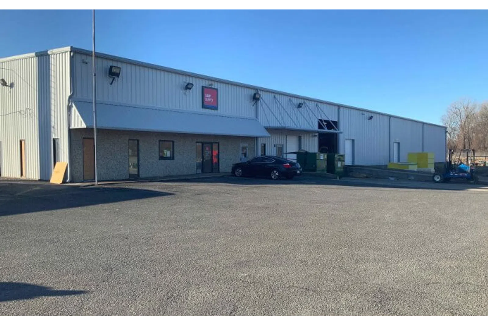 Large industrial warehouse with metal siding under clear blue sky and a parked black car outside.