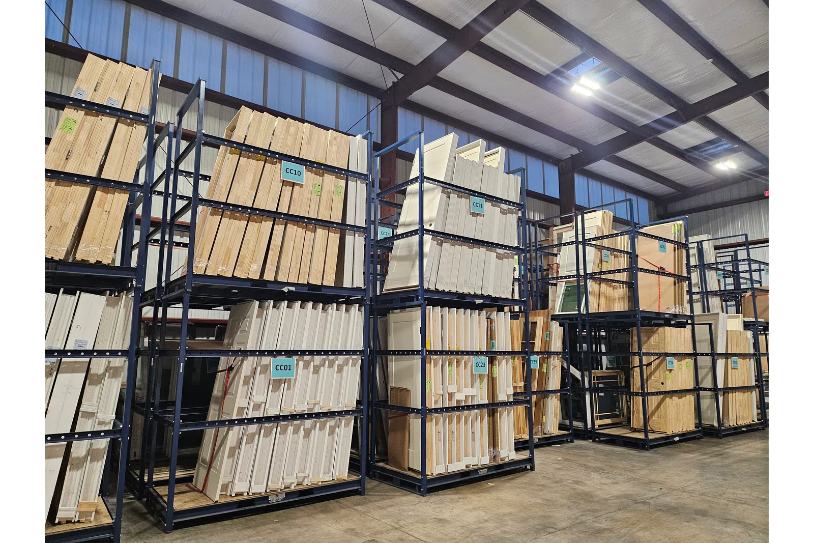Wood panels and lumber stacked on tall metal racks in a spacious warehouse with concrete floors and high ceilings.