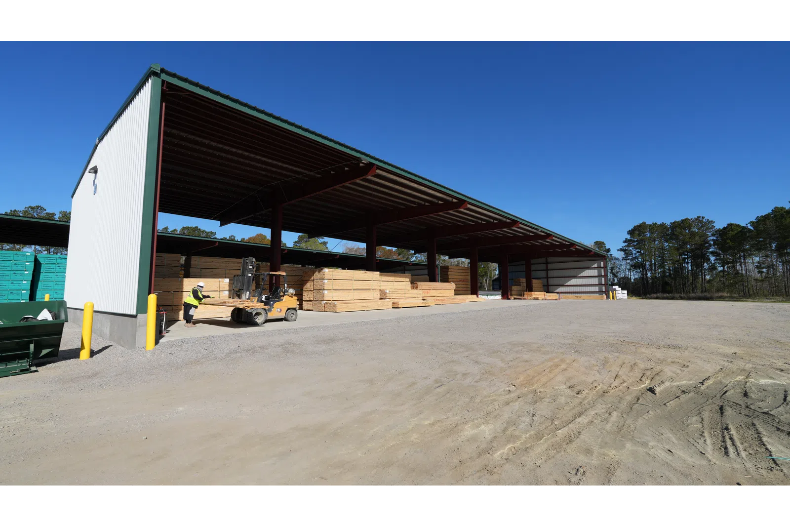 Covered outdoor storage facility with stacked lumber and a forklift operator under clear blue sky.